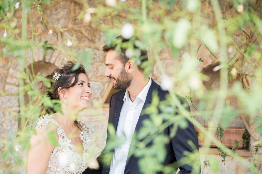 Romantic bride and groom portrait framed by soft greenery, captured against a rustic stone wall in a sun-washed Mediterranean setting. Their warm smiles and tender gaze create an intimate, timeless moment—perfect for destination wedding photography in a charming historic village.