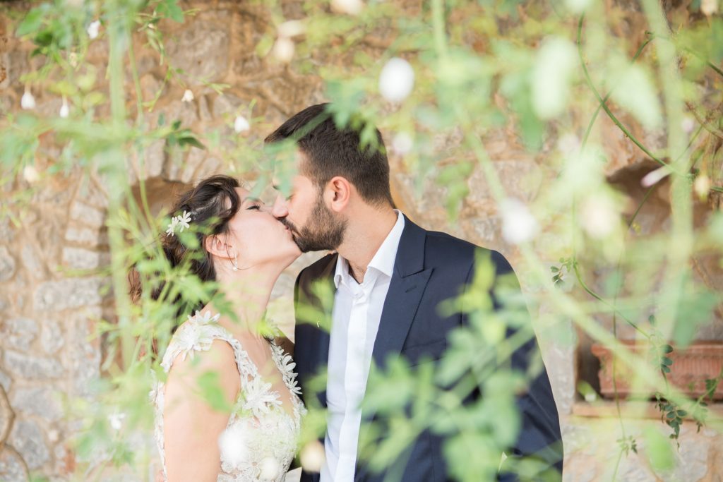 Romantic bride and groom portrait framed by soft greenery, captured against a rustic stone wall in a sun-washed Mediterranean setting. Their warm smiles and tender gaze create an intimate, timeless moment—perfect for destination wedding photography in a charming historic village.