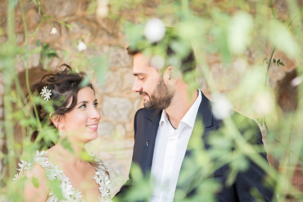 Romantic bride and groom portrait framed by soft greenery, captured against a rustic stone wall in a sun-washed Mediterranean setting. Their warm smiles and tender gaze create an intimate, timeless moment—perfect for destination wedding photography in a charming historic village.
