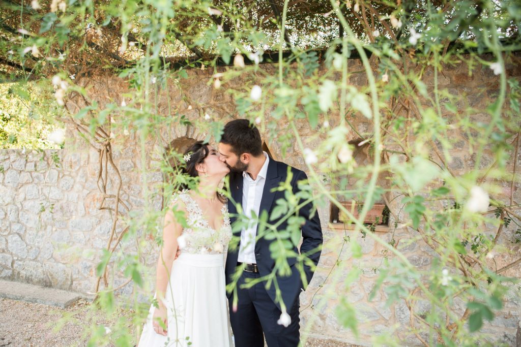 Romantic bride and groom portrait framed by soft greenery, captured against a rustic stone wall in a sun-washed Mediterranean setting. Their warm smiles and tender gaze create an intimate, timeless moment—perfect for destination wedding photography in a charming historic village.