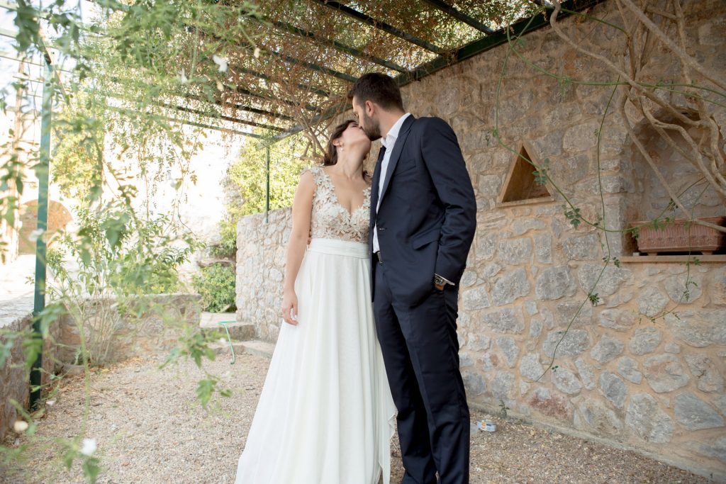 Romantic bride and groom portrait framed by soft greenery, captured against a rustic stone wall in a sun-washed Mediterranean setting. Their warm smiles and tender gaze create an intimate, timeless moment—perfect for destination wedding photography in a charming historic village.