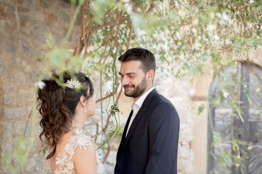 Romantic bride and groom portrait framed by soft greenery, captured against a rustic stone wall in a sun-washed Mediterranean setting. Their warm smiles and tender gaze create an intimate, timeless moment—perfect for destination wedding photography in a charming historic village.
