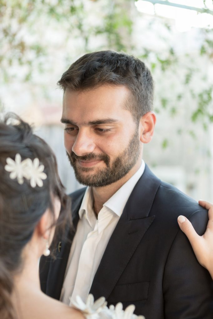 Romantic bride and groom portrait framed by soft greenery, captured against a rustic stone wall in a sun-washed Mediterranean setting. Their warm smiles and tender gaze create an intimate, timeless moment—perfect for destination wedding photography in a charming historic village.