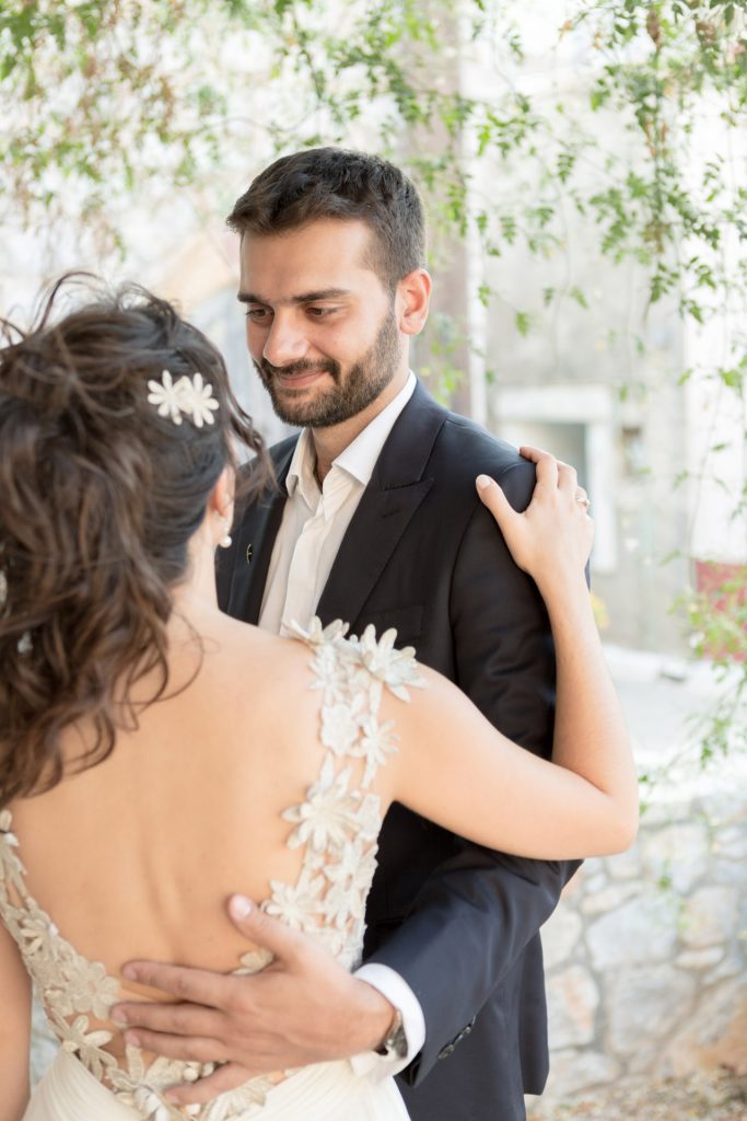 Romantic bride and groom portrait framed by soft greenery, captured against a rustic stone wall in a sun-washed Mediterranean setting. Their warm smiles and tender gaze create an intimate, timeless moment—perfect for destination wedding photography in a charming historic village.