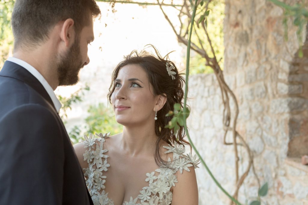 Romantic bride and groom portrait framed by soft greenery, captured against a rustic stone wall in a sun-washed Mediterranean setting. Their warm smiles and tender gaze create an intimate, timeless moment—perfect for destination wedding photography in a charming historic village.