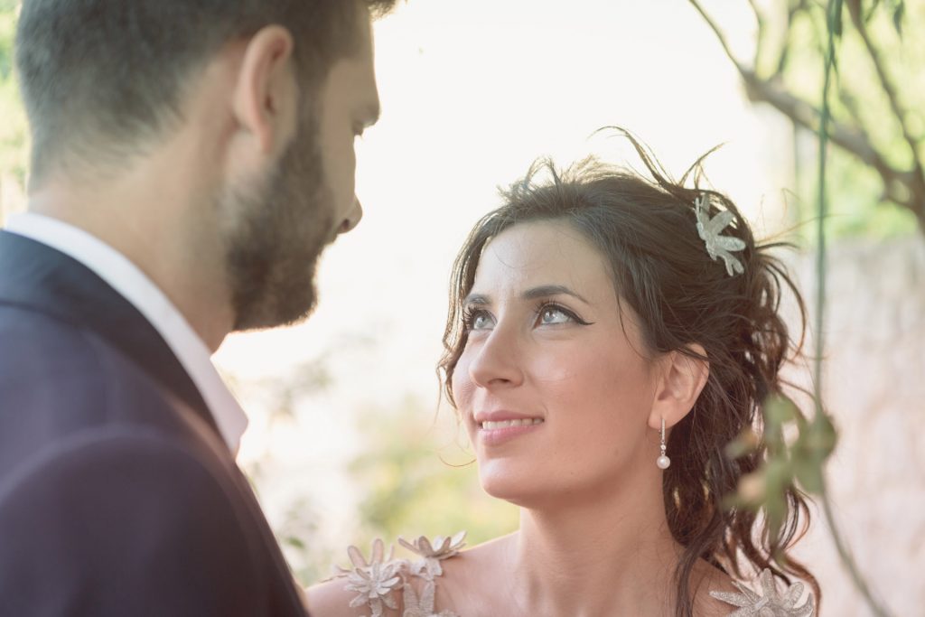 Romantic bride and groom portrait framed by soft greenery, captured against a rustic stone wall in a sun-washed Mediterranean setting. Their warm smiles and tender gaze create an intimate, timeless moment—perfect for destination wedding photography in a charming historic village.