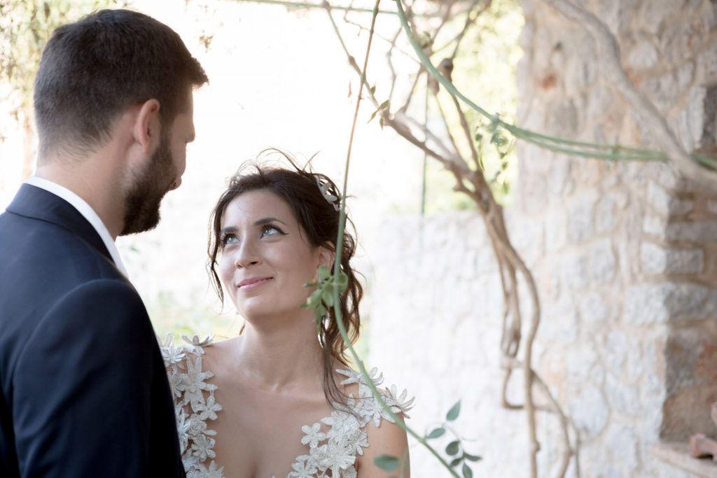 Romantic bride and groom portrait framed by soft greenery, captured against a rustic stone wall in a sun-washed Mediterranean setting. Their warm smiles and tender gaze create an intimate, timeless moment—perfect for destination wedding photography in a charming historic village.