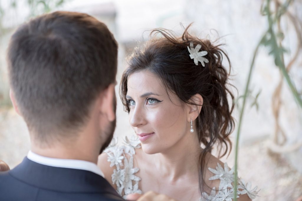 Romantic bride and groom portrait framed by soft greenery, captured against a rustic stone wall in a sun-washed Mediterranean setting. Their warm smiles and tender gaze create an intimate, timeless moment—perfect for destination wedding photography in a charming historic village.