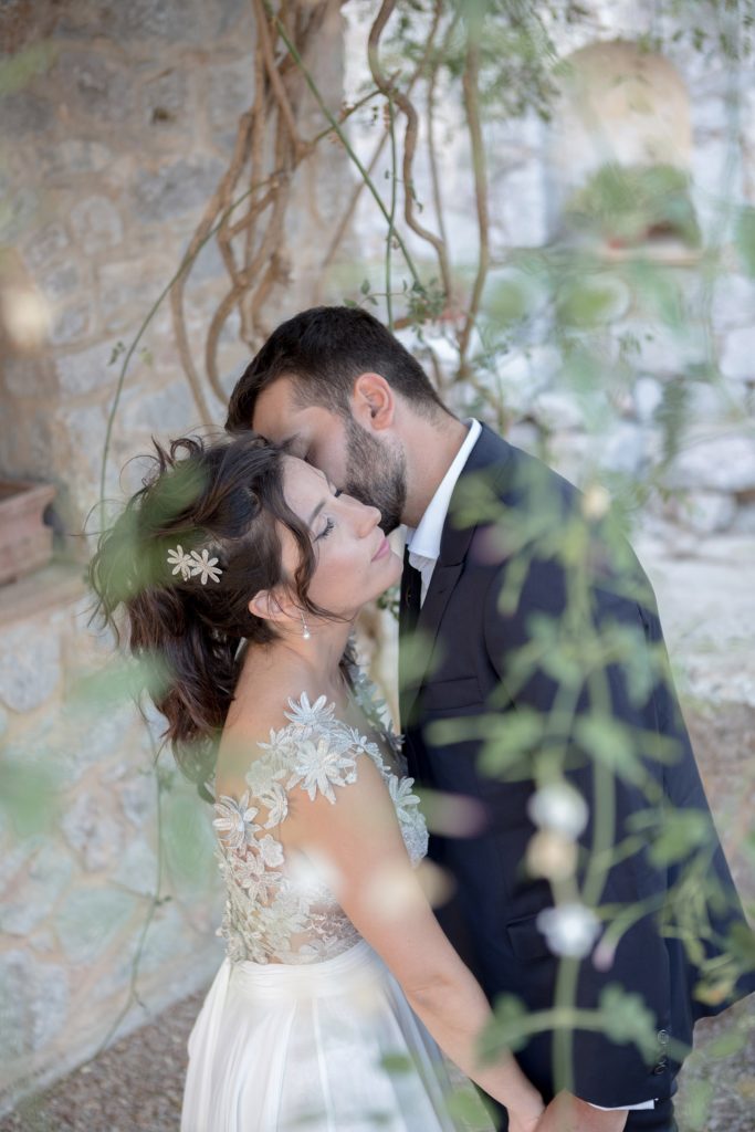 Romantic bride and groom portrait framed by soft greenery, captured against a rustic stone wall in a sun-washed Mediterranean setting. Their warm smiles and tender gaze create an intimate, timeless moment—perfect for destination wedding photography in a charming historic village.