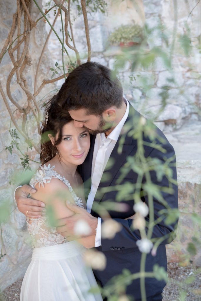 Romantic bride and groom portrait framed by soft greenery, captured against a rustic stone wall in a sun-washed Mediterranean setting. Their warm smiles and tender gaze create an intimate, timeless moment—perfect for destination wedding photography in a charming historic village.