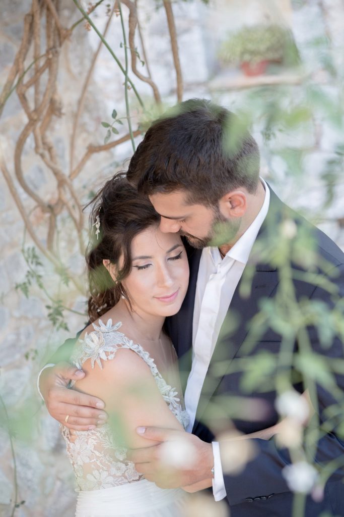 Romantic bride and groom portrait framed by soft greenery, captured against a rustic stone wall in a sun-washed Mediterranean setting. Their warm smiles and tender gaze create an intimate, timeless moment—perfect for destination wedding photography in a charming historic village.