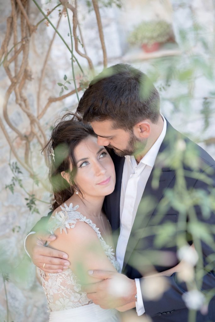 Romantic bride and groom portrait framed by soft greenery, captured against a rustic stone wall in a sun-washed Mediterranean setting. Their warm smiles and tender gaze create an intimate, timeless moment—perfect for destination wedding photography in a charming historic village.