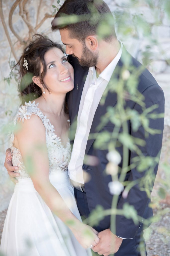 Romantic bride and groom portrait framed by soft greenery, captured against a rustic stone wall in a sun-washed Mediterranean setting. Their warm smiles and tender gaze create an intimate, timeless moment—perfect for destination wedding photography in a charming historic village.