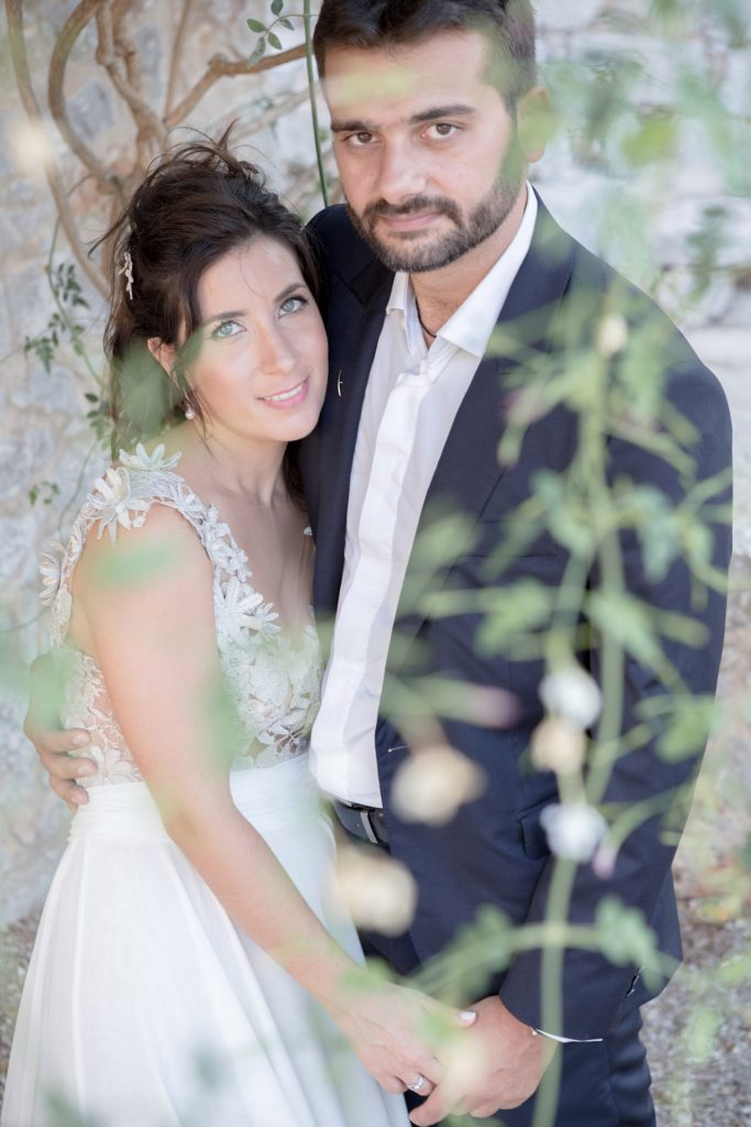 Romantic bride and groom portrait framed by soft greenery, captured against a rustic stone wall in a sun-washed Mediterranean setting. Their warm smiles and tender gaze create an intimate, timeless moment—perfect for destination wedding photography in a charming historic village.