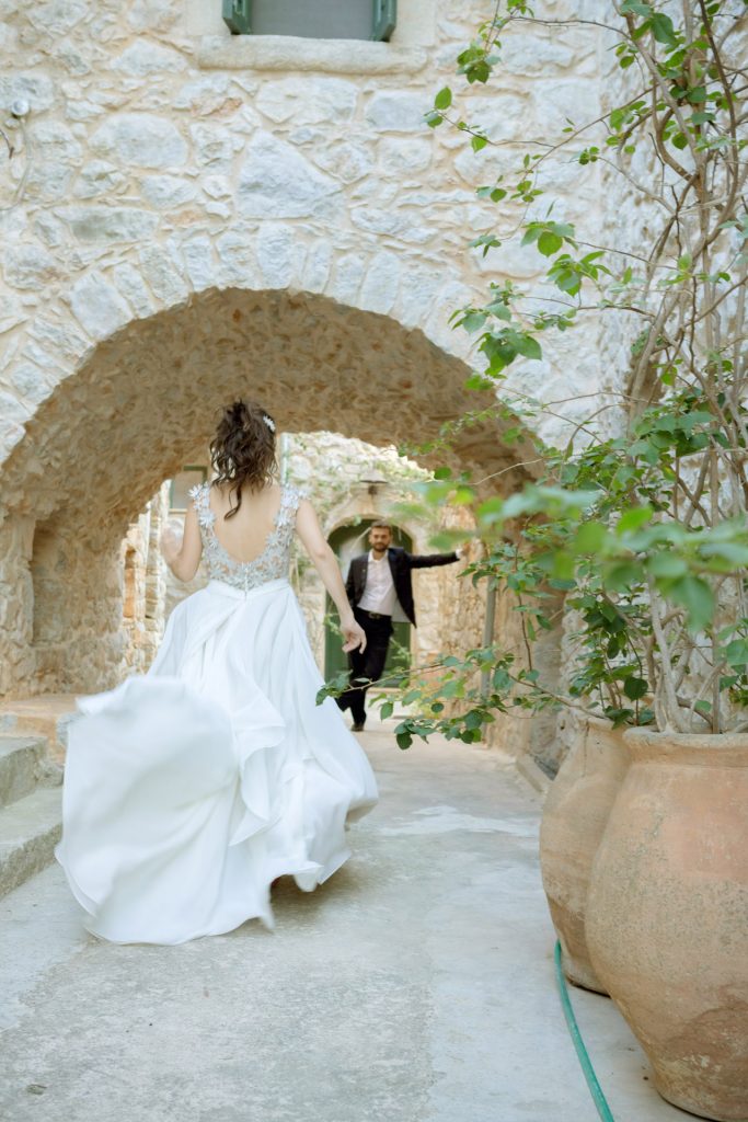 Romantic bride and groom portrait framed by soft greenery, captured against a rustic stone wall in a sun-washed Mediterranean setting. Their warm smiles and tender gaze create an intimate, timeless moment—perfect for destination wedding photography in a charming historic village.