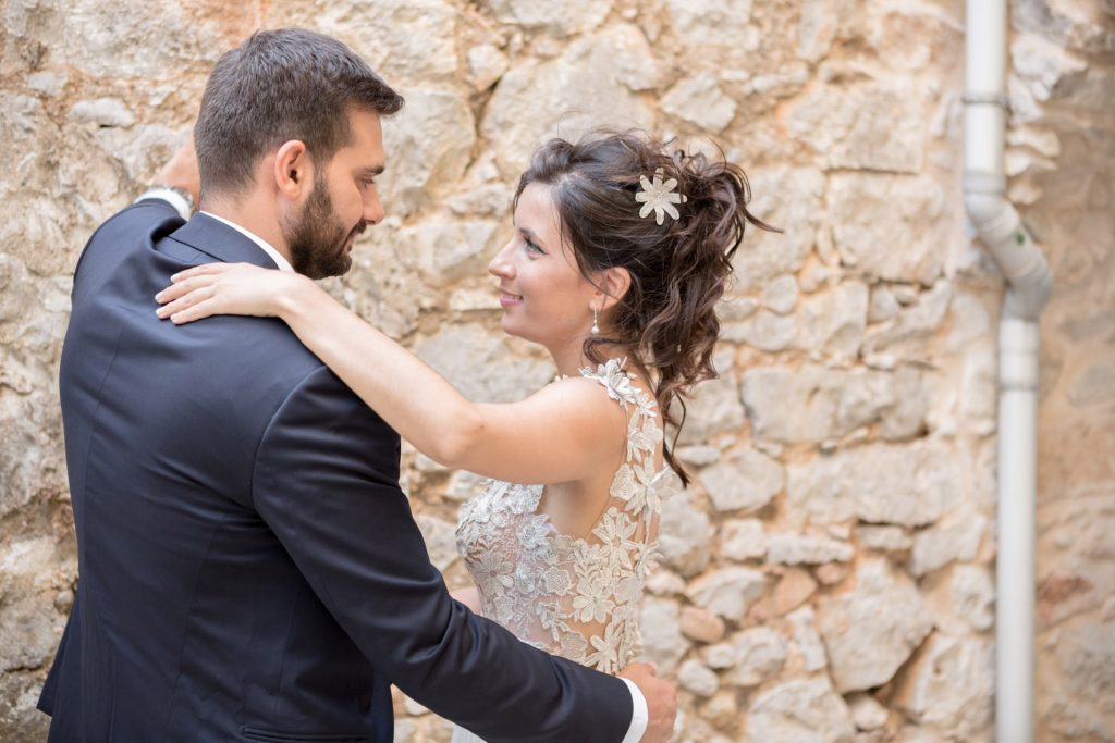Romantic bride and groom portrait framed by soft greenery, captured against a rustic stone wall in a sun-washed Mediterranean setting. Their warm smiles and tender gaze create an intimate, timeless moment—perfect for destination wedding photography in a charming historic village.