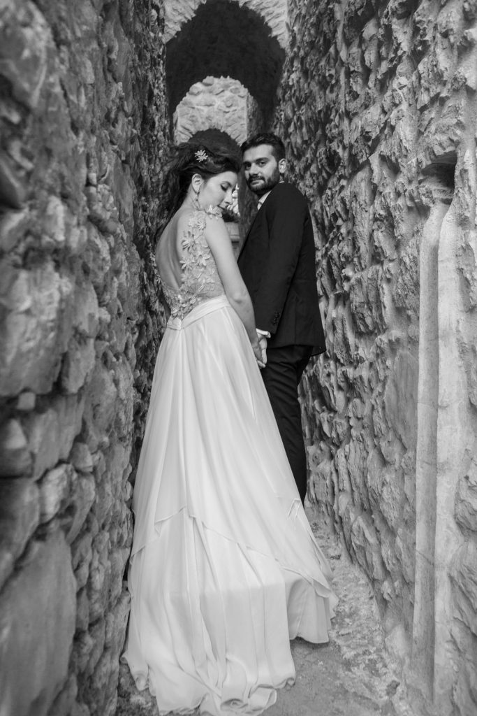Romantic bride and groom b&w portrait framed by soft greenery, captured against a rustic stone wall in a sun-washed Mediterranean setting. Their warm smiles and tender gaze create an intimate, timeless moment—perfect for destination wedding photography in a charming historic village.