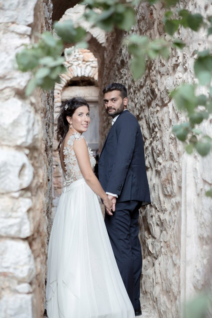 Romantic bride and groom portrait framed by soft greenery, captured against a rustic stone wall in a sun-washed Mediterranean setting. Their warm smiles and tender gaze create an intimate, timeless moment—perfect for destination wedding photography in a charming historic village.
