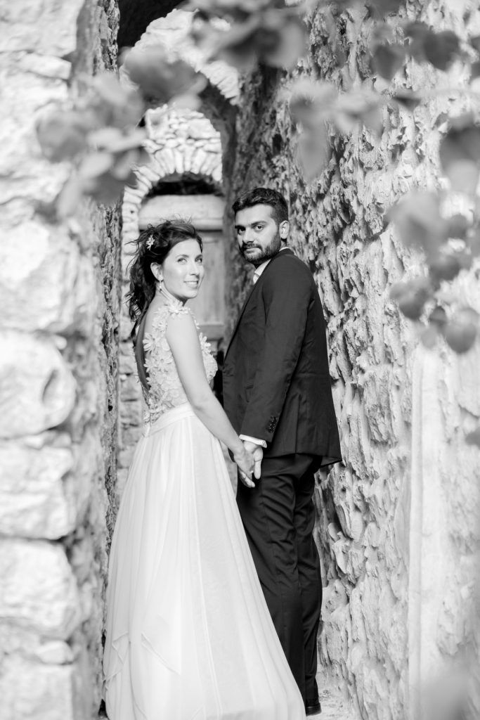 Romantic bride and groom b&w portrait framed by soft greenery, captured against a rustic stone wall in a sun-washed Mediterranean setting. Their warm smiles and tender gaze create an intimate, timeless moment—perfect for destination wedding photography in a charming historic village.