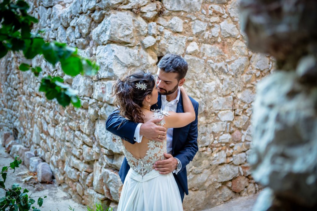 Romantic bride and groom portrait framed by soft greenery, captured against a rustic stone wall in a sun-washed Mediterranean setting. Their warm smiles and tender gaze create an intimate, timeless moment—perfect for destination wedding photography in a charming historic village.
