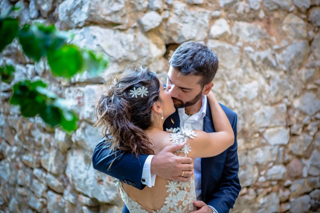 Romantic bride and groom portrait framed by soft greenery, captured against a rustic stone wall in a sun-washed Mediterranean setting. Their warm smiles and tender gaze create an intimate, timeless moment—perfect for destination wedding photography in a charming historic village.