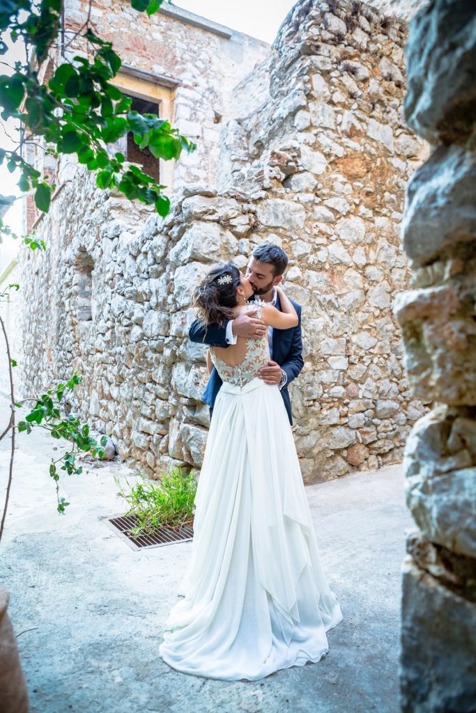 Romantic bride and groom portrait framed by soft greenery, captured against a rustic stone wall in a sun-washed Mediterranean setting. Their warm smiles and tender gaze create an intimate, timeless moment—perfect for destination wedding photography in a charming historic village.
