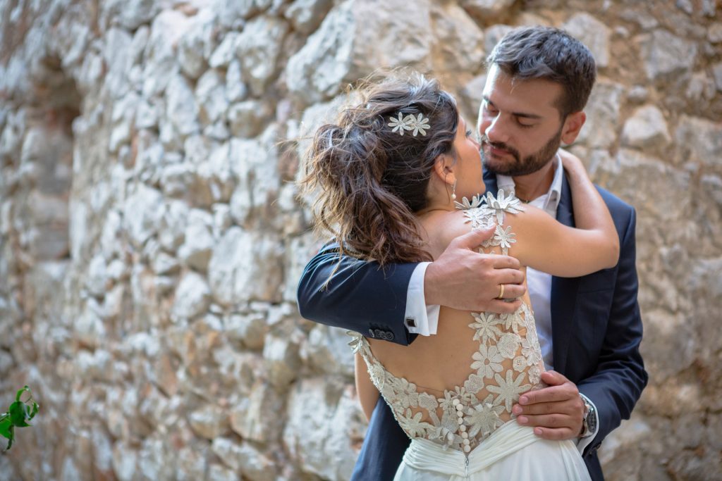 Romantic bride and groom portrait framed by soft greenery, captured against a rustic stone wall in a sun-washed Mediterranean setting. Their warm smiles and tender gaze create an intimate, timeless moment—perfect for destination wedding photography in a charming historic village.