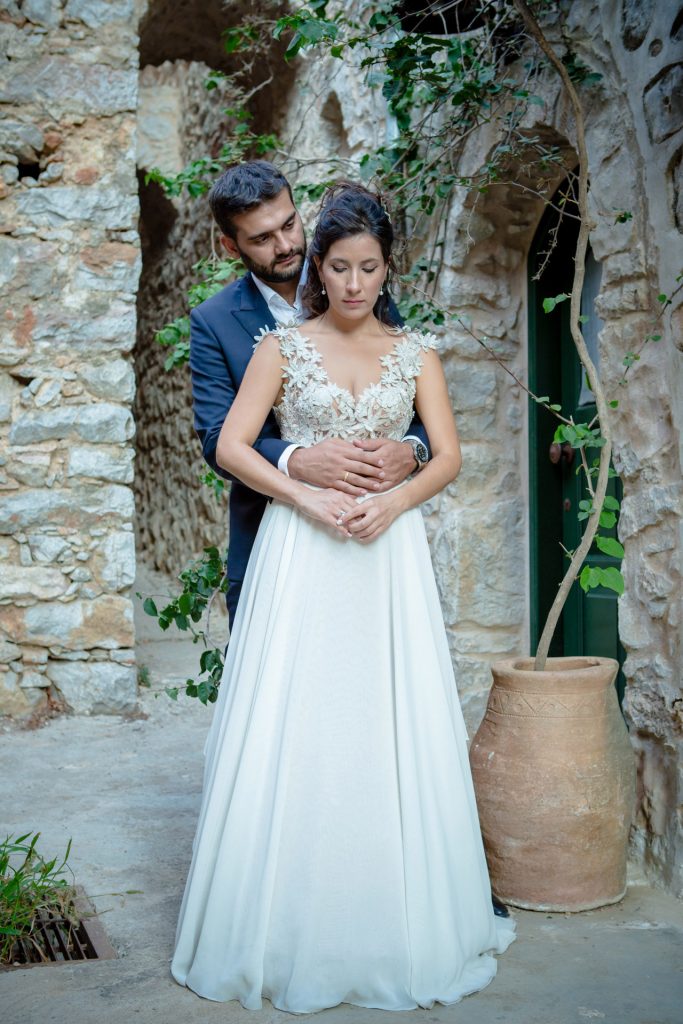 Romantic bride and groom portrait framed by soft greenery, captured against a rustic stone wall in a sun-washed Mediterranean setting. Their warm smiles and tender gaze create an intimate, timeless moment—perfect for destination wedding photography in a charming historic village.