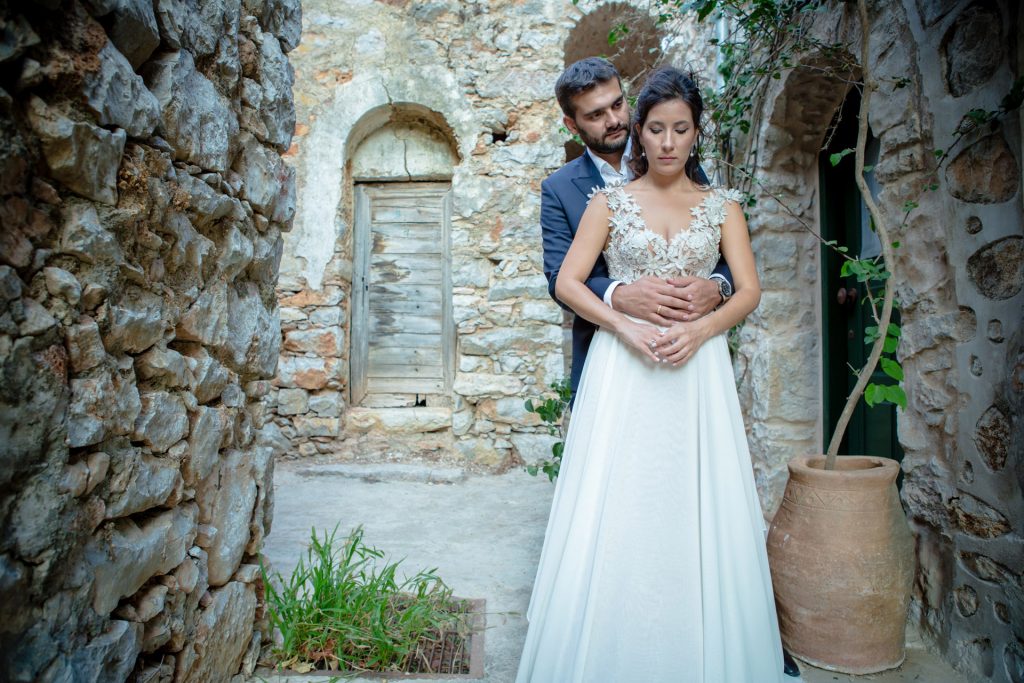 Romantic bride and groom portrait framed by soft greenery, captured against a rustic stone wall in a sun-washed Mediterranean setting. Their warm smiles and tender gaze create an intimate, timeless moment—perfect for destination wedding photography in a charming historic village.