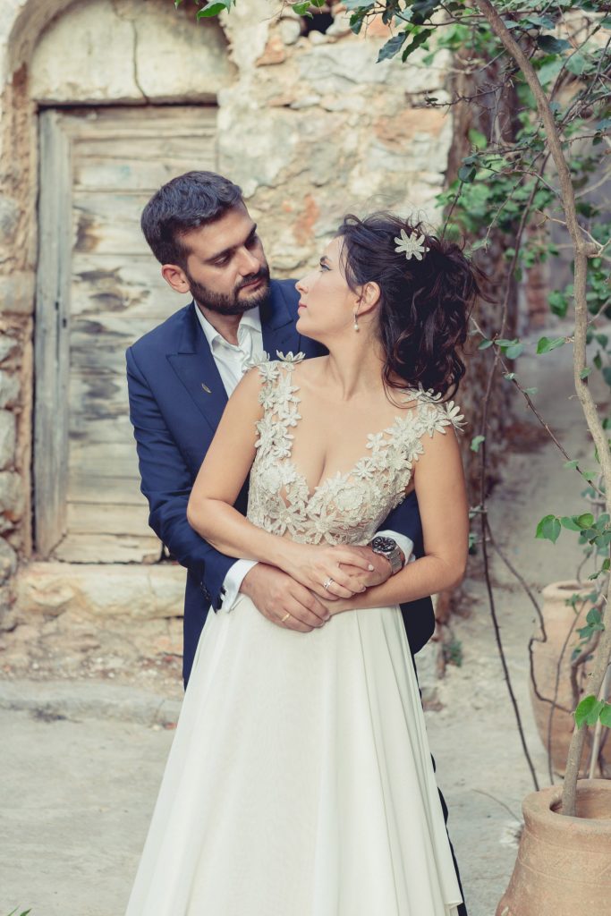 Romantic bride and groom portrait framed by soft greenery, captured against a rustic stone wall in a sun-washed Mediterranean setting. Their warm smiles and tender gaze create an intimate, timeless moment—perfect for destination wedding photography in a charming historic village.
