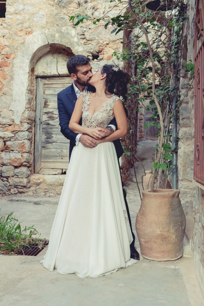 Romantic bride and groom portrait framed by soft greenery, captured against a rustic stone wall in a sun-washed Mediterranean setting. Their warm smiles and tender gaze create an intimate, timeless moment—perfect for destination wedding photography in a charming historic village.