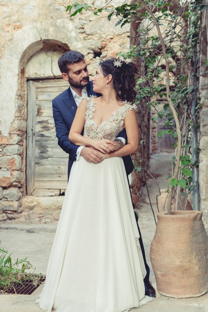 Romantic bride and groom portrait framed by soft greenery, captured against a rustic stone wall in a sun-washed Mediterranean setting. Their warm smiles and tender gaze create an intimate, timeless moment—perfect for destination wedding photography in a charming historic village.