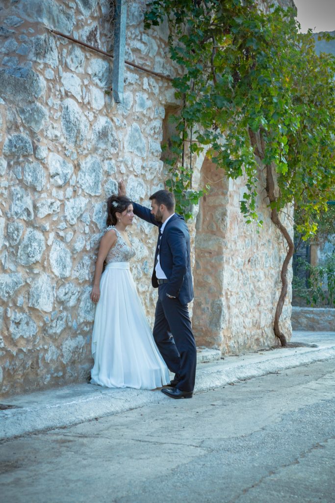 Romantic bride and groom portrait framed by soft greenery, captured against a rustic stone wall in a sun-washed Mediterranean setting. Their warm smiles and tender gaze create an intimate, timeless moment—perfect for destination wedding photography in a charming historic village.