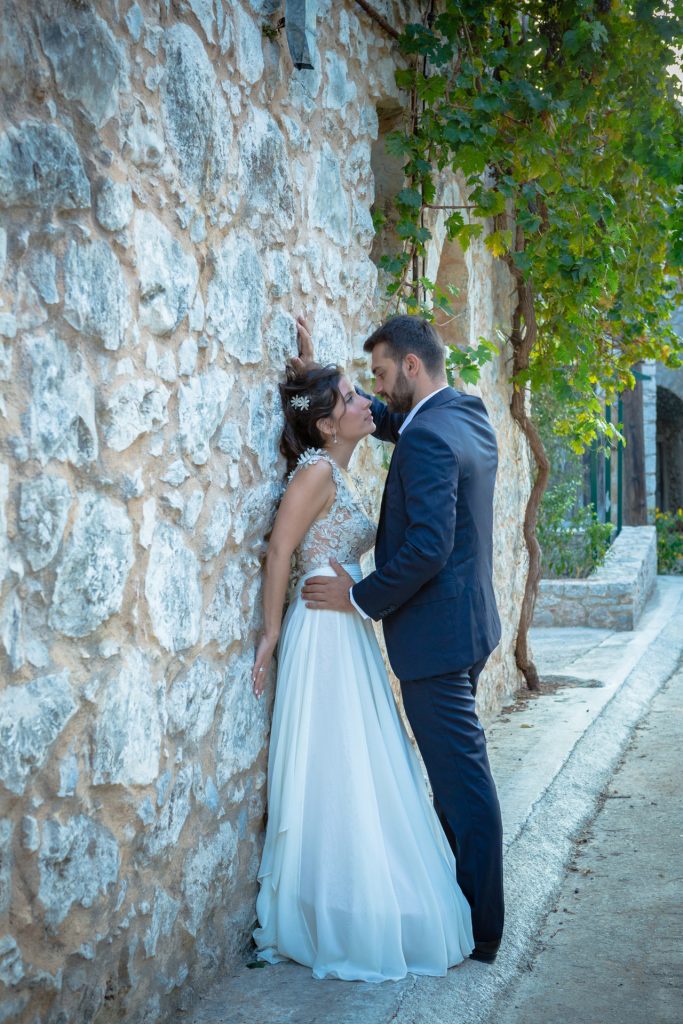 Romantic bride and groom portrait framed by soft greenery, captured against a rustic stone wall in a sun-washed Mediterranean setting. Their warm smiles and tender gaze create an intimate, timeless moment—perfect for destination wedding photography in a charming historic village.
