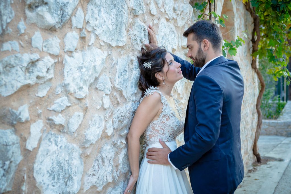 Romantic bride and groom portrait framed by soft greenery, captured against a rustic stone wall in a sun-washed Mediterranean setting. Their warm smiles and tender gaze create an intimate, timeless moment—perfect for destination wedding photography in a charming historic village.