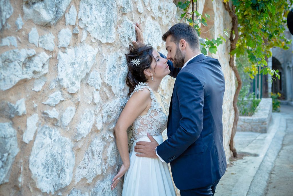 Romantic bride and groom portrait framed by soft greenery, captured against a rustic stone wall in a sun-washed Mediterranean setting. Their warm smiles and tender gaze create an intimate, timeless moment—perfect for destination wedding photography in a charming historic village.