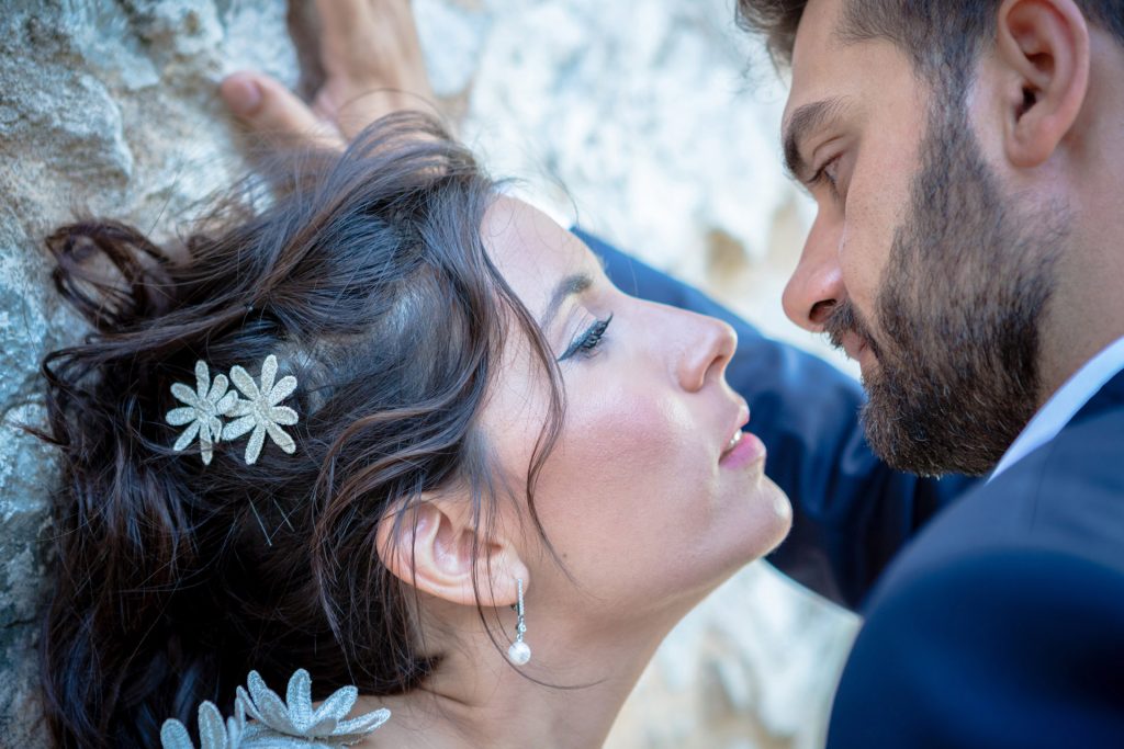 Romantic bride and groom portrait framed by soft greenery, captured against a rustic stone wall in a sun-washed Mediterranean setting. Their warm smiles and tender gaze create an intimate, timeless moment—perfect for destination wedding photography in a charming historic village.