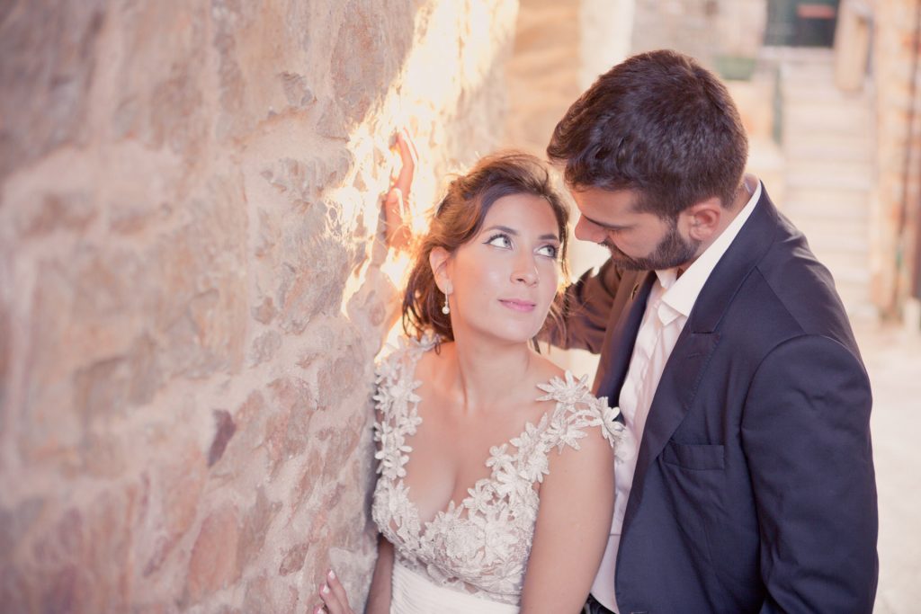 Romantic bride and groom portrait framed by soft greenery, captured against a rustic stone wall in a sun-washed Mediterranean setting. Their warm smiles and tender gaze create an intimate, timeless moment—perfect for destination wedding photography in a charming historic village.