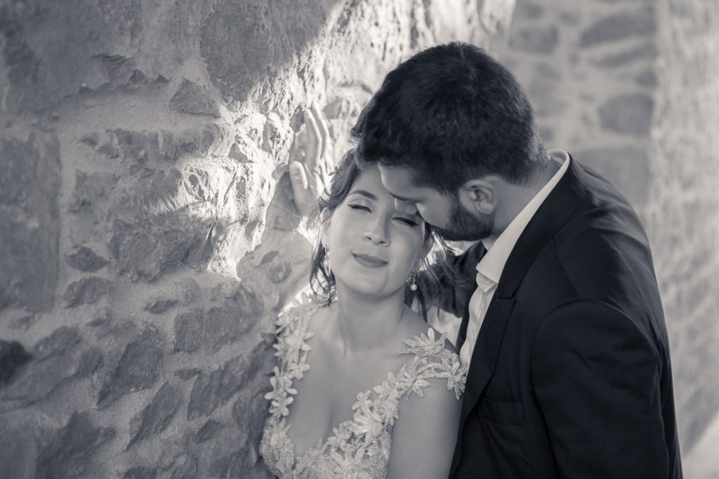 Romantic bride and groom portrait framed by soft greenery, captured against a rustic stone wall in a sun-washed Mediterranean setting. Their warm smiles and tender gaze create an intimate, timeless moment—perfect for destination wedding photography in a charming historic village.