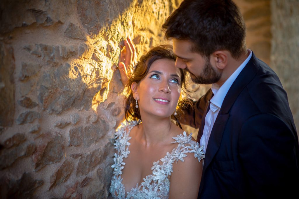 Romantic bride and groom portrait framed by soft greenery, captured against a rustic stone wall in a sun-washed Mediterranean setting. Their warm smiles and tender gaze create an intimate, timeless moment—perfect for destination wedding photography in a charming historic village.